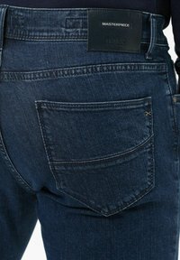 Jeans in denim scuro con taglio dritto, cuciture visibili, una tasca posteriore singola e un'etichetta in pelle con logo in vita.