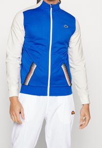 Blå och grå zip-jacka med texturerade sidopaneler med ett geometriskt mönster, en rund logotyp på vänster bröst och ribbade fållar.