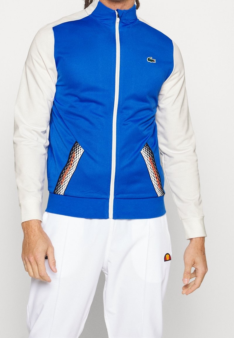 Blå och grå zip-jacka med texturerade sidopaneler med ett geometriskt mönster, en rund logotyp på vänster bröst och ribbade fållar.