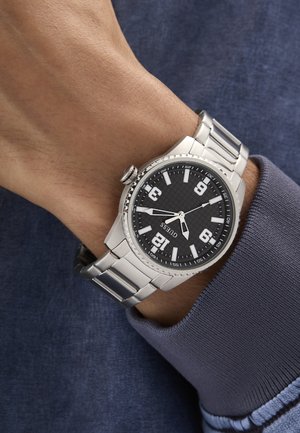 Zapešće s satom na srebrnoj metalnoj narukvici koji pokazuje vrijeme 8:20, crni brojčanik s bijelim brojevima i oznakama, nošen preko plave manžete dugih rukava.