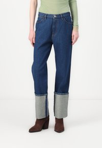 Modre kavbojke z udobnim krojem, ki imajo sivo obrobo in tradicionalen gumbasti pas. Oblečene so z rjavimi čevlji na peto.