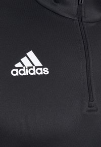 Czarna sportowa bluzka na suwak, wykonana z gładkiego, teksturowanego materiału. Wyróżnia się białym logo adidas po lewej stronie klatki piersiowej.