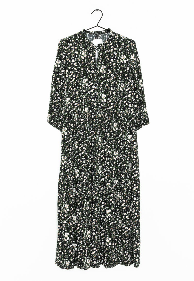 Vestido negro con estampado floral blanco, mangas 3/4, escote en V, cintura fruncida, largo hasta los tobillos, textura fluida, dobladillo ondulado. Cuelga de un gancho.
