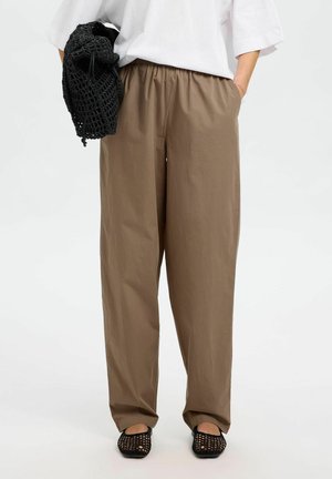 Losse, hoge bruine broeken met een elastische tailleband, zijzakken en lichte stof. Gecombineerd met een zwarte netten tas en platte schoenen.
