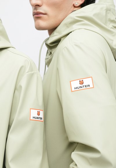Δύο άτομα φορούν ελαφριά μπεζ αδιάβροχα Hunter με λογότυπα κεφαλής αλεπούς σε πορτοκαλί χρώμα στα μανίκια, το πρόσωπο ενός ατόμου είναι μερικώς ορατό.