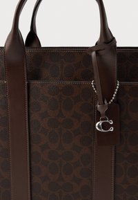 Brun läderväska med ett mönstrat yta, slät textur, dubbla handtag och en metalltagg som har en logotypcharms.