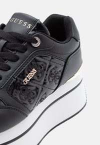 Black sneaker with patent leather upper, embossed logo, and metallic accents. Features a thick white sole and black laces.