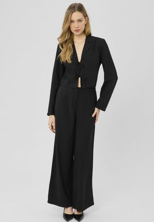 Femme aux longs cheveux blonds portant un blazer court noir à liens avant, un pantalon noir à jambes larges, des talons noirs pointus et un collier délicat.