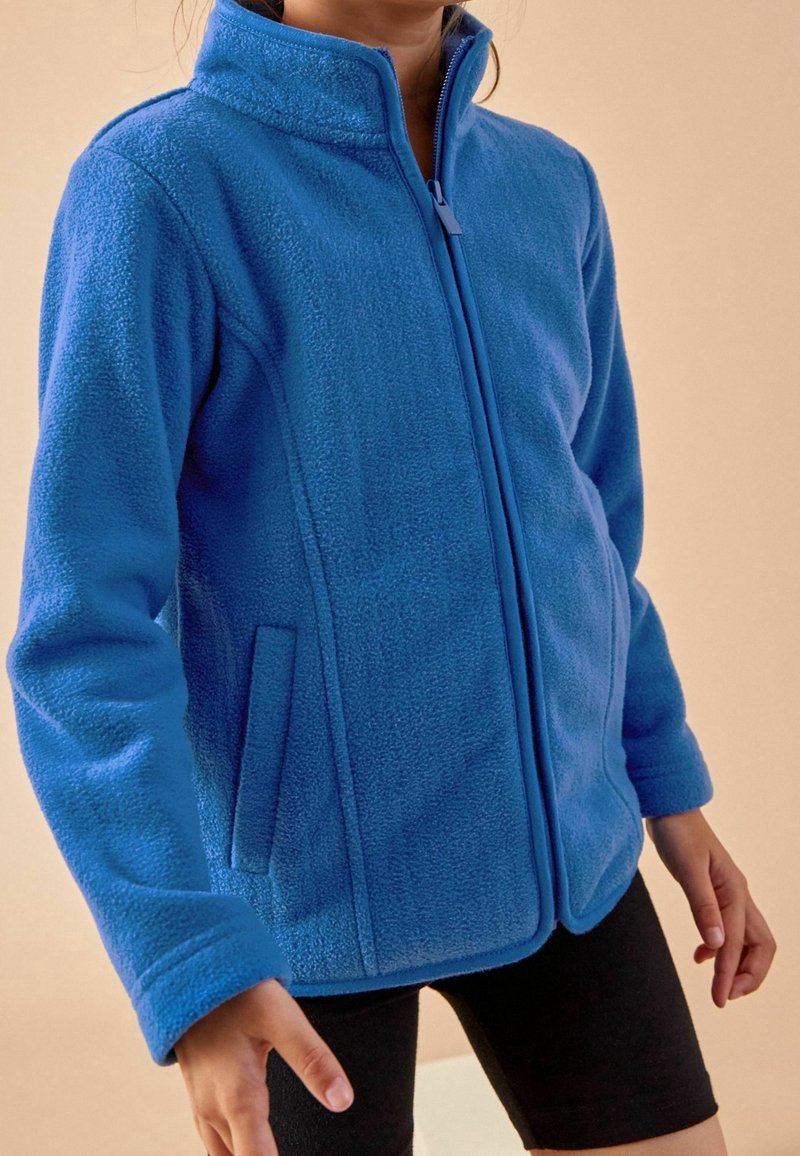 Personne portant une veste polaire zippée bleu vif avec poches latérales et un short noir sur un fond beige uni.