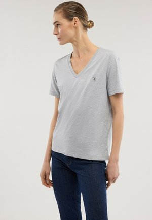 Frau mit zusammengebundenem Haar in einem hellgrauen V-Ausschnitt-T-Shirt mit kurzen Ärmeln und dunkelblauen Jeans, schaut nach links vor schlichtem Hintergrund.