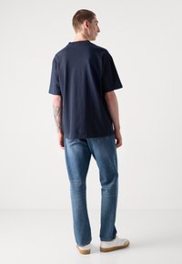 Námořnická modrá košile s krátkým rukávem a volným střihem, nošená s světle modrými džíny a bílými botami. Má kulatý výstřih a rovný lem.