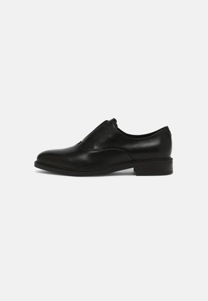 Chaussure de ville en cuir noir, à enfiler, avec un faible talon, finition lisse et détails de couture minimalistes sur fond blanc.