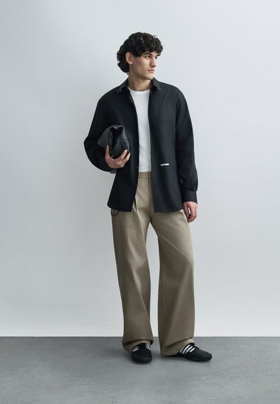 Chemise noire boutonnée portée sur un t-shirt blanc, pantalon beige à jambes larges, et baskets noires à rayures blanches. Tient une pochette noire.