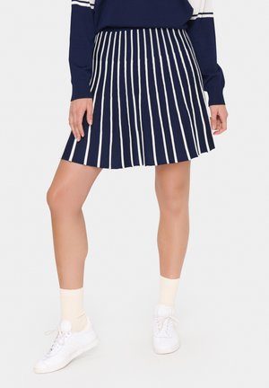 Marineblauwe plisseerok met verticale witte strepen, gemaakt van een zachte stof. Gecombineerd met witte sneakers en crèmekleurige enkelkousen.