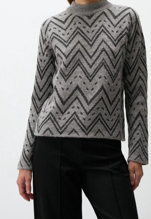 Personne portant un pull gris avec un motif zigzag et à pois noirs, assorti à un pantalon noir, debout devant un fond uni clair.