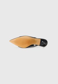 Chaussure noire à bout pointu avec un design ouvert, surface lisse et semelle en caoutchouc beige présentant des rainures horizontales pour une meilleure adhérence. Détail du logo doré.