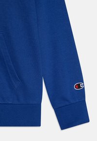 Sudadera azul con tejido texturizado, bolsillo frontal y puños acanalados. Logo bordado notable en la manga izquierda.