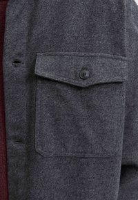 Camisa de mezcla de lana en gris oscuro, con un acabado texturizado, que presenta un bolsillo en el pecho con botón y detalle de botón negro.