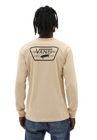Mann mit Dreadlocks, der ein beiges Langarm-Vans-Shirt mit schwarzem Logo und Skateboard-Grafik auf dem Rücken trägt, kombiniert mit blauen Jeans.