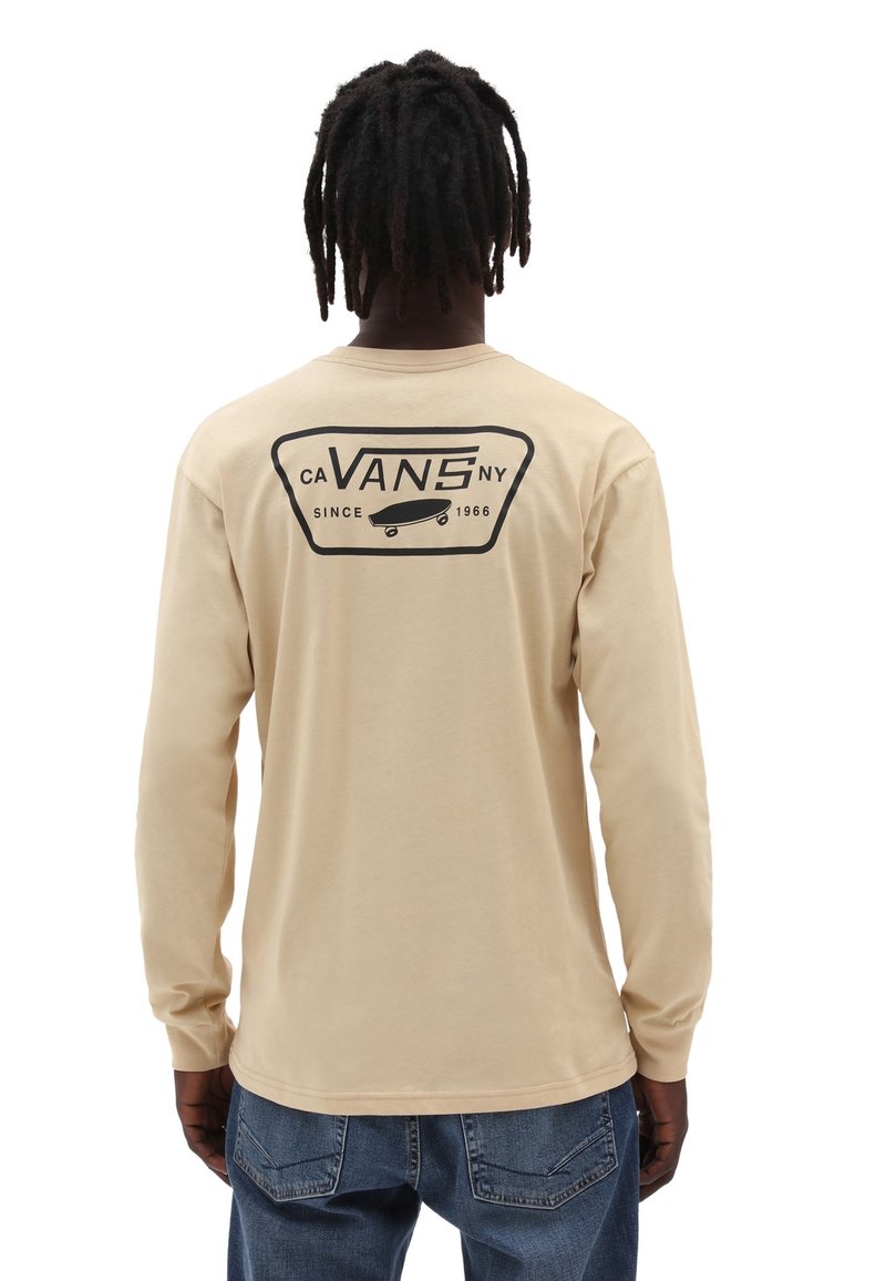 Mann mit Dreadlocks, der ein beiges Langarm-Vans-Shirt mit schwarzem Logo und Skateboard-Grafik auf dem Rücken trägt, kombiniert mit blauen Jeans.