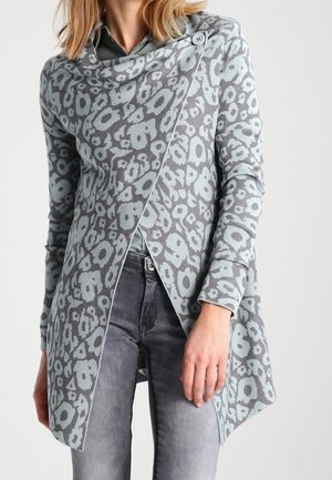 Femme portant un cardigan asymétrique imprimé léopard gris et bleu clair sur un t-shirt gris et un jean gris délavé.