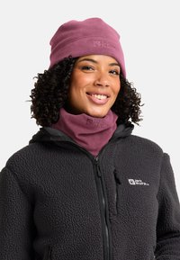 Donna sorridente con capelli ricci che indossa un cappello in pile rosa e un scaldacollo, oltre a una giacca in pile nera con zip e logo sul lato sinistro del petto.