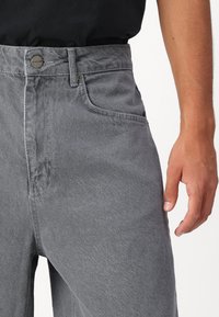 Pantalon en denim gris à coupe droite, présentant une fermeture à bouton, des poches avant et une texture subtile. Porté avec une chemise noire.