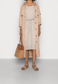 Beige lange trenchcoat over een jurk met bloempatroon. Model draagt een tan kleurige mand tas en heeft tan kleurige platte sandalen aan. Glad, lichtgewicht texturen.