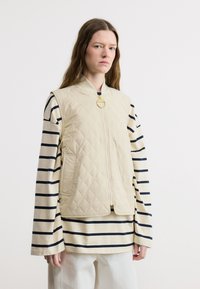 Γυναίκα που φοράει μπεζ γιλέκο χωρίς μανίκια με καπιτονέ σχέδιο πάνω από μια μακρυμάνικη κρεμ μπλούζα με μαύρες οριζόντιες ρίγες και λευκό παντελόνι, στέκεται μπροστά σε λευκό φόντο.