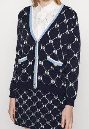 Cardigan bleu marine et jupe avec motif géométrique blanc, bordure bleu clair, boutons dorés, portés sur un chemisier blanc à col.