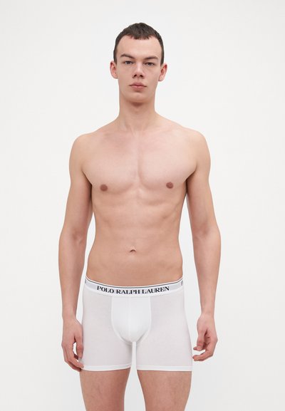 Witte katoenen boxershorts met een zwarte logo tailleband, aansluitend design, gladde textuur en lengte tot halverwege de dij. Model staat, zonder shirt.