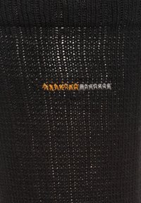 Chaussette noire texturée avec des côtes verticales, ornée de coutures horizontales en orange et gris près du centre.