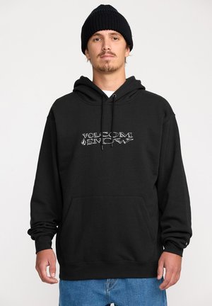 Hoodie noir en tissu épais, avec une poche kangourou à l'avant et un logo emblématique en argent. À associer avec un pantalon en denim bleu.