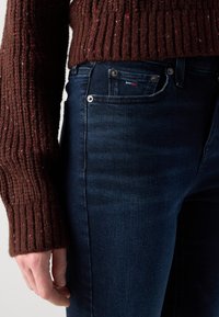Dark blue jeans with a slim fit, featuring a small front pocket, brass rivets, and a textured brown knitted sweater with red specks.