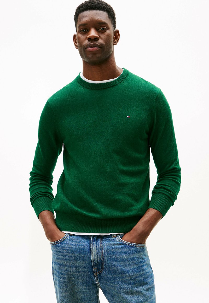 Mand iført en grøn crewneck-sweater over en hvid skjorte, med hænderne i lommerne på blå jeans, stående mod en ensfarvet hvid baggrund.