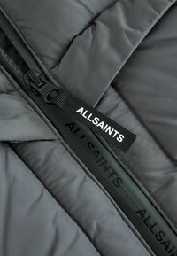 Grå puffjacka med kvadra textur, utrustad med en svart dragkedja och en vävd etikett med texten "ALLSAINTS."