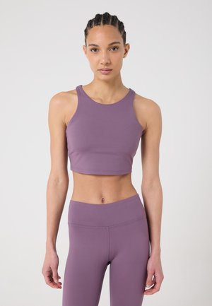 Mujer con el cabello trenzado, vestida con un top corto sin mangas color malva y leggings a juego de cintura alta, de pie contra un fondo claro y liso.