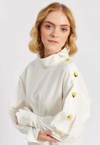 Blond naine naine lühikeste laineliste juustega, kes kannab valget pluusi, millel on kuldsed nööbid õlgadel ja varrukatel, käed õrnalt ristis.