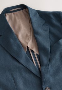 Blauer Leinenblazer mit Reverskragen, teilweise geöffnet und enthüllt ein hellbeiges, gemustertes Futter. Verfügt über einen Knopfverschluss und eine dezente Textur.