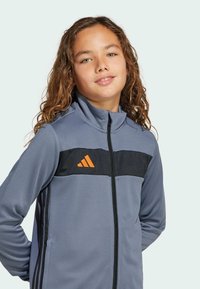 Chaqueta deportiva gris con cremallera, panel negro y logotipo naranja. Presenta un alto cuello, cierre de cremallera y bolsillos laterales. Textura suave.