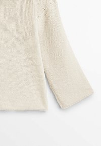 Manche de pull tricotée couleur crème avec un large poignet et un motif de point texturé et épais sur un fond blanc.