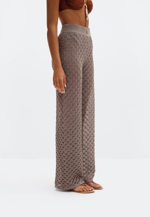 Kvinne iført høyt liv, vide, teksturerte strikkebukser i taupe med beige midjebånd og brune thongsandaler mot hvit bakgrunn.