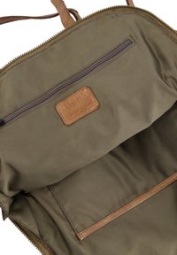 Olive green canvas bag interior with a brown leather logo patch, a zip pocket, and a smooth texture. Features a zipper closure.