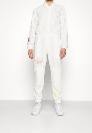 Persoon in een witte jumpsuit met kleurrijke geborduurde abstracte figuren, Adidas-logo op de linkerbovenbeen en witte Adidas-sneakers.