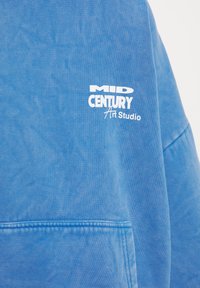 Felpa azzurra chiara con tessuto testurizzato, dotata di tasca frontale e logo stampato bianco con la scritta "MID CENTURY Art Studio."