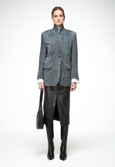 Mujer con blazer gris de cuello alto, falda midi de cuero negra con abertura frontal, botas negras hasta la rodilla, sosteniendo un bolso negro.