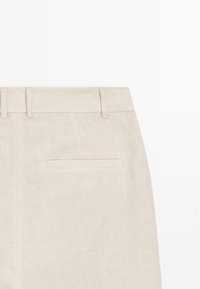 Pantalon en lin beige avec passants pour ceinture et une poche passepoilée unique à l’arrière, présenté sur un fond blanc uni.