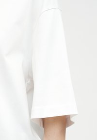 Valkoinen lyhythihainen paita päällä, jossa näkyy hihan sauman yksityiskohta ja osa käsivarresta yksiväristä taustaa vasten.