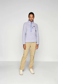 Lichtpaarse fleece pullover met een opstaande kraag, ritssluiting en contrasterende blauwe accenten. Gecombineerd met beige broeken en lichte schoenen.