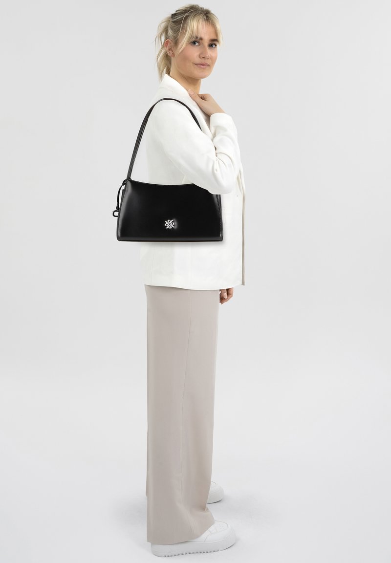 Sac à bandoulière en cuir noir au design épuré, forme arrondie et accent de logo argenté au centre. Le modèle porte un manteau blanc et un pantalon beige.